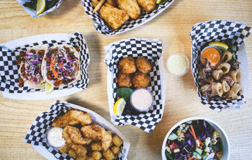 A variety of dishes on a wooden table, including tacos, fried fish, and sides, all in checkered paper baskets.