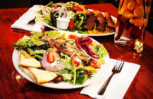 Two plates of fresh salad with grilled chicken, tomatoes, onions, and pita bread, served on a wooden table.