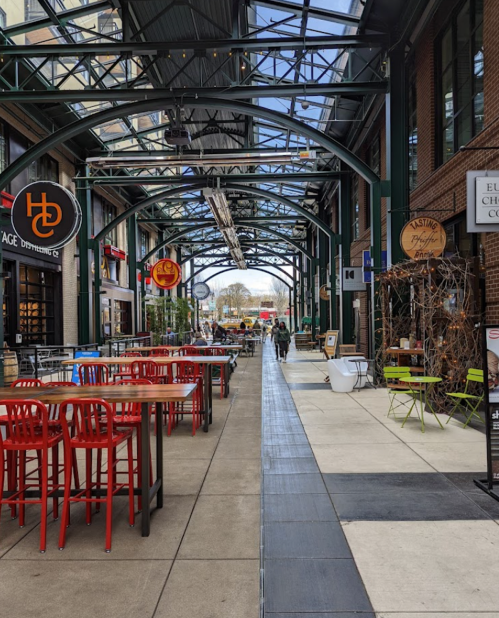 A vibrant indoor market with red tables, green chairs, and a spacious walkway under a glass roof.
