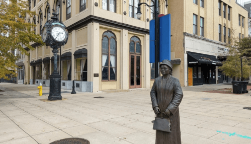 A bronze statue of a woman stands near a vintage clock on a city street with shops and autumn trees in the background.