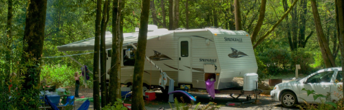 A camper trailer parked in a wooded area, surrounded by trees and outdoor gear.