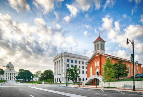 A scenic view of a street with historic buildings, including a red brick church and a white government building, under a cloudy sky.