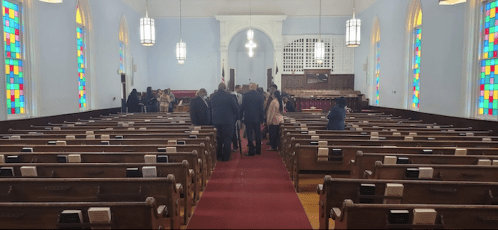 Interior of a church with colorful stained glass windows and people gathered in the aisle. Rows of wooden pews visible.