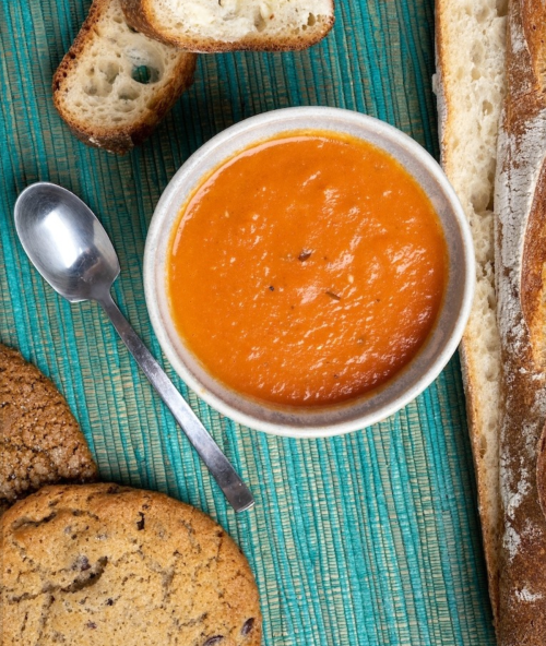 A bowl of tomato soup with a spoon, surrounded by slices of bread and chocolate chip cookies on a textured surface.