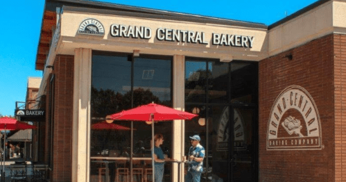 A bakery storefront with a sign reading "Grand Central Bakery," featuring outdoor seating and two people chatting.