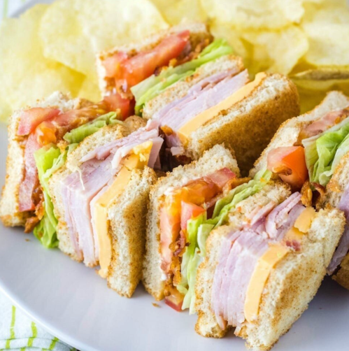 A close-up of a layered sandwich with ham, cheese, lettuce, and tomato, served with a side of potato chips.
