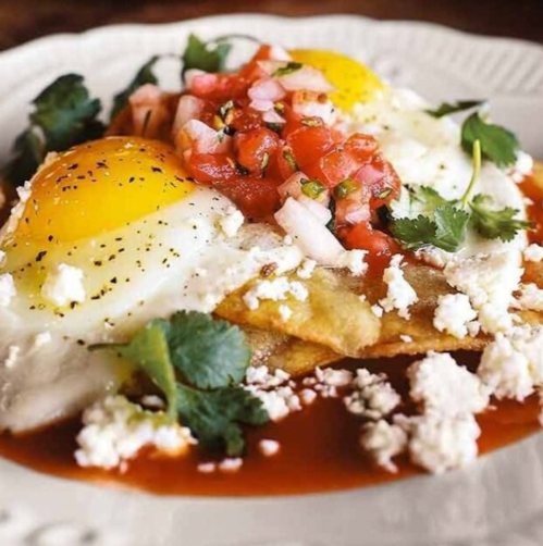 A plate of huevos rancheros with sunny-side-up eggs, salsa, cilantro, and crumbled cheese on tortillas.