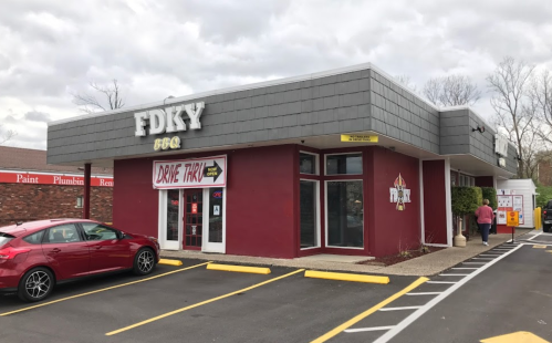Exterior of a BBQ restaurant with a drive-thru, featuring a red and gray facade and parked cars in the lot.
