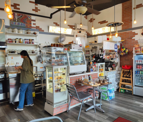 A cozy café interior with a counter, display cases of pastries, and a person preparing food in the kitchen area.