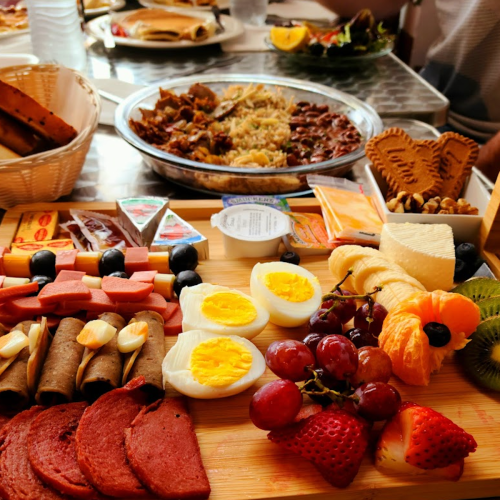 A vibrant spread of assorted cheeses, fruits, meats, and snacks on a wooden platter, with a large dish of rice in the background.
