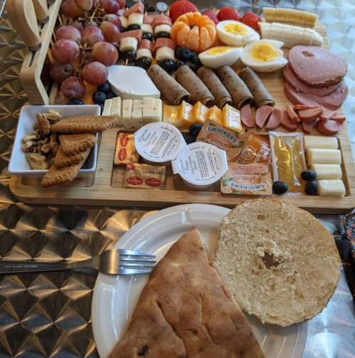 A charcuterie board with meats, cheeses, fruits, and snacks, alongside a plate with a bagel and a fork.