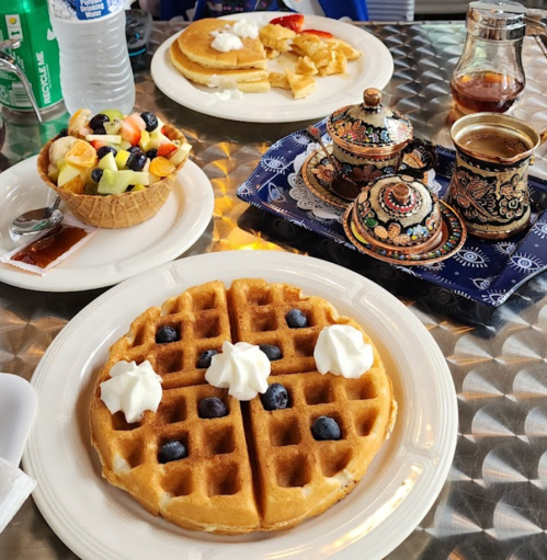 A table with a plate of waffles topped with whipped cream and blueberries, alongside pancakes, fruit, and tea.