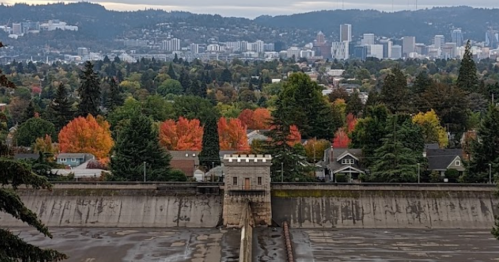 View of a dam with a city skyline in the background, surrounded by autumn foliage and overcast skies.