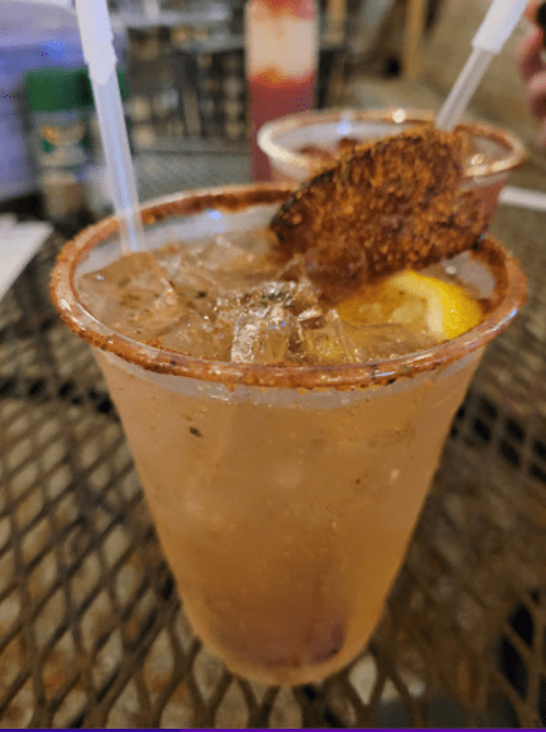 Two drinks with ice, garnished with a slice of fruit and a rim of seasoning, on a metal table.