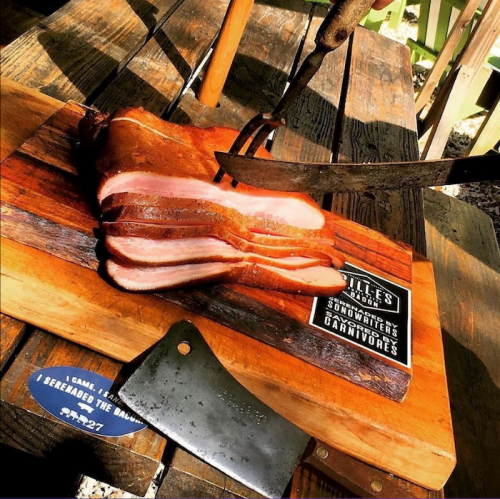 A wooden cutting board with sliced bacon and a cleaver, set outdoors with greenery in the background.