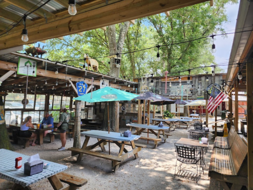 Outdoor dining area with picnic tables, umbrellas, and string lights, surrounded by trees and a rustic backdrop.