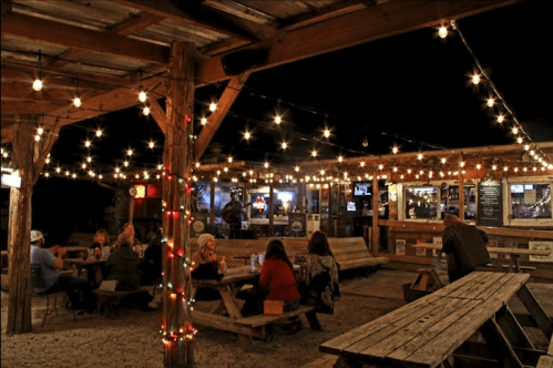 A cozy outdoor venue at night, with string lights illuminating wooden tables and people enjoying drinks and music.