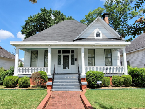 A charming white house with a front porch, surrounded by green bushes and a brick pathway, under a clear blue sky.