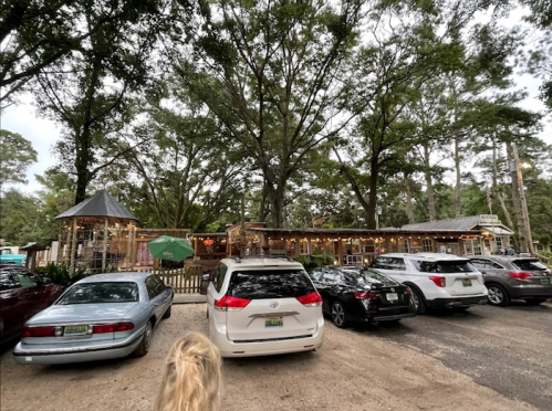 A rustic restaurant surrounded by trees, with several parked cars in the foreground and outdoor seating visible.