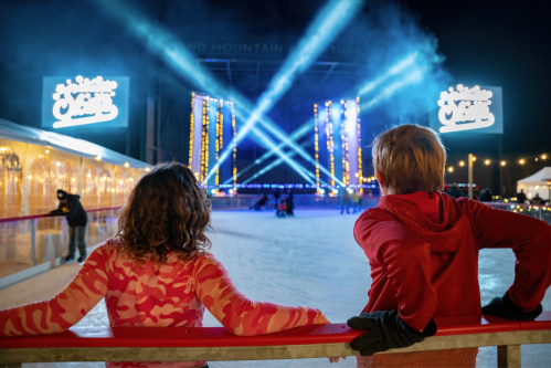 Two children watch a festive ice skating rink illuminated by colorful lights and a large sign in the background.