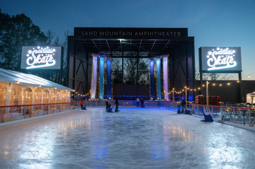 An outdoor ice skating rink at dusk, with festive lights and a large sign reading "Skate" in the background.
