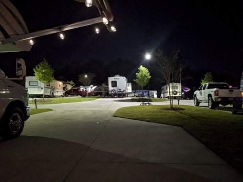 A nighttime scene of a campground with RVs and parked vehicles illuminated by string lights and a street lamp.