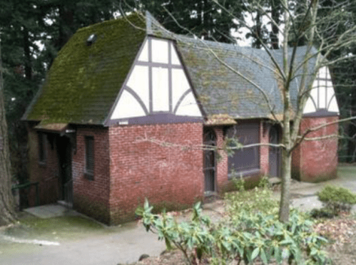 A small, rustic brick building with a sloped roof and moss, surrounded by trees and greenery.