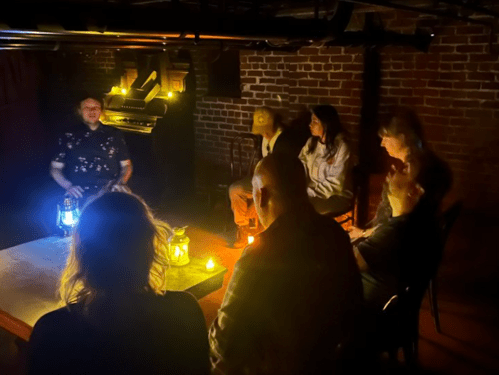 A dimly lit room with a group of people seated around a table, listening to a speaker holding a lantern.