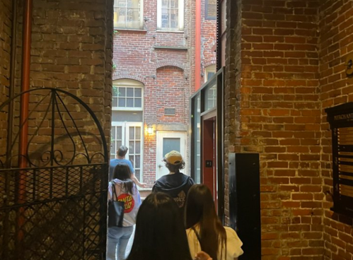 A group of people walks through a brick hallway towards an open door leading to a courtyard.