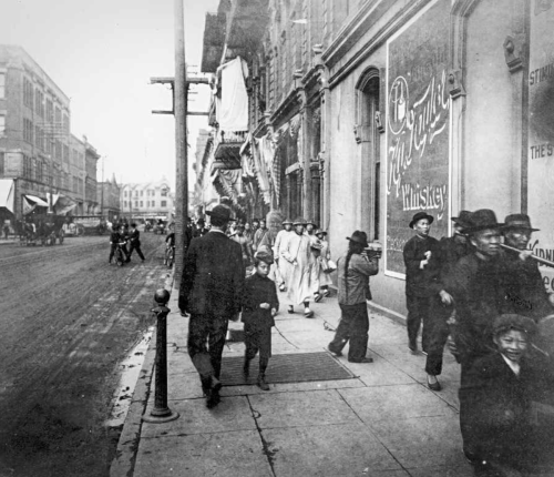 Black and white photo of a busy street scene with pedestrians, horse-drawn carriages, and storefronts from the early 1900s.