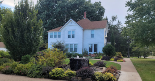 A charming two-story white house surrounded by lush greenery and colorful landscaping, with a small fountain in the garden.