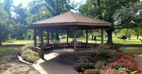 A wooden gazebo surrounded by colorful flowers in a sunny park setting.