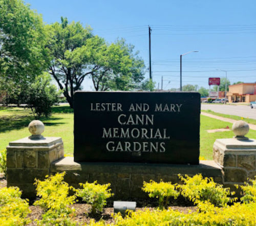 Sign for Lester and Mary Cann Memorial Gardens, surrounded by greenery and flowers on a sunny day.