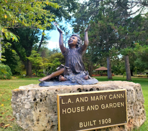 Bronze statue of a joyful girl with arms raised, beside a plaque for the L.A. and Mary Cann House and Garden, built 1908.
