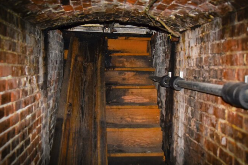 A dimly lit staircase leading up through a brick-walled basement, with wooden steps and beams visible.