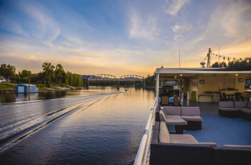 A scenic view of a river at sunset, with a boat cruising and a bridge in the background. Comfortable seating on the deck.