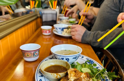 A table with bowls of food and people using chopsticks to eat, surrounded by vibrant decor.