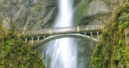 A scenic waterfall cascades down rocky cliffs, with a stone bridge arching over the water below, surrounded by lush greenery.