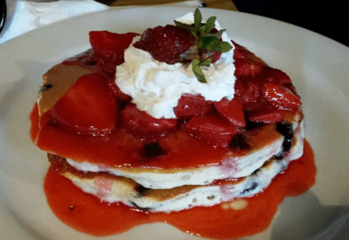 Stack of pancakes topped with strawberries, whipped cream, and strawberry sauce on a white plate.