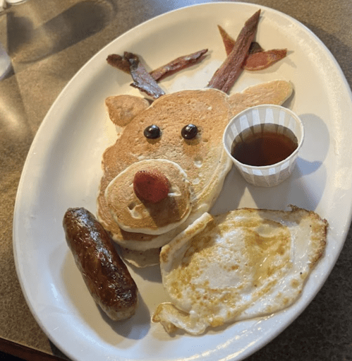 A playful breakfast plate featuring pancakes shaped like a reindeer, with bacon antlers, sausage, and an egg.