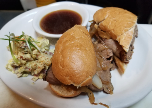 Two sandwiches with beef and onions on a plate, accompanied by a small bowl of dipping sauce and a side of coleslaw.