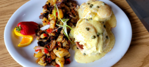 A plate of eggs Benedict with hollandaise sauce, served with seasoned hash browns and fresh fruit on the side.