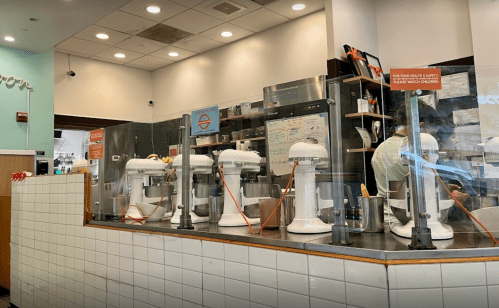A bakery counter with several white mixers and a menu board in the background, featuring a clean, modern design.