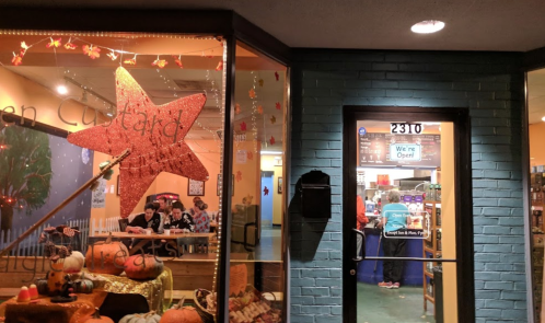 A cozy storefront with a large star decoration, pumpkins outside, and people dining inside. Sign reads "We're Open!"