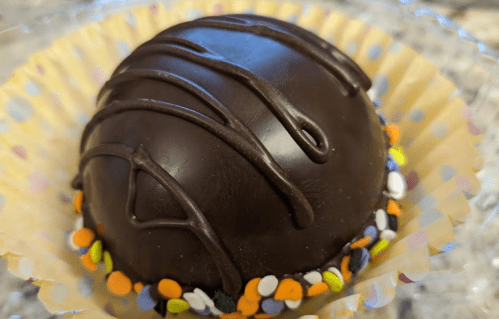 A round chocolate treat topped with drizzled chocolate and colorful sprinkles on a cupcake liner.
