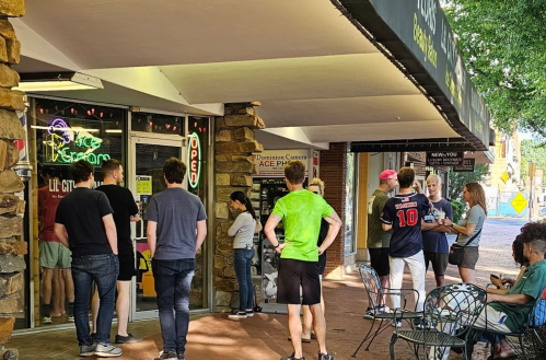 A group of people stands outside an ice cream shop, chatting and waiting in a lively street scene.
