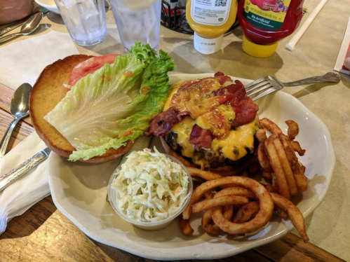 A plate featuring a cheeseburger with bacon, lettuce, tomato, curly fries, and a side of coleslaw.