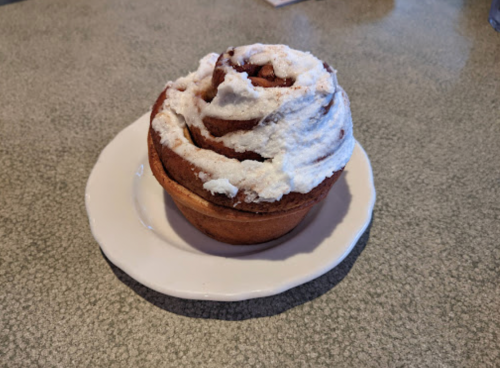 A freshly baked cinnamon roll topped with fluffy white frosting on a small white plate.