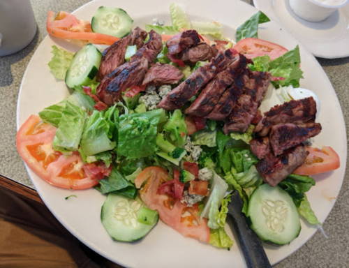 A fresh salad topped with grilled steak, tomatoes, cucumbers, and mixed greens on a white plate.