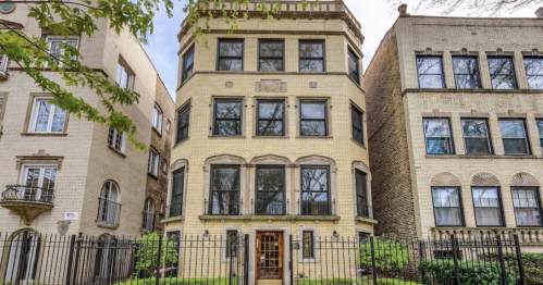 A three-story brick building with large windows, surrounded by greenery and a black iron fence.